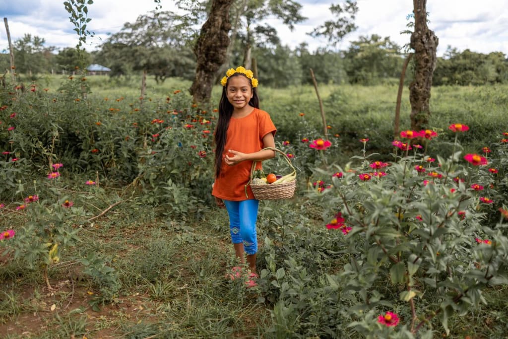 Maira is wearing an orange shirt with jeans. She is holding a basket full of vegetables and standing in a garden.